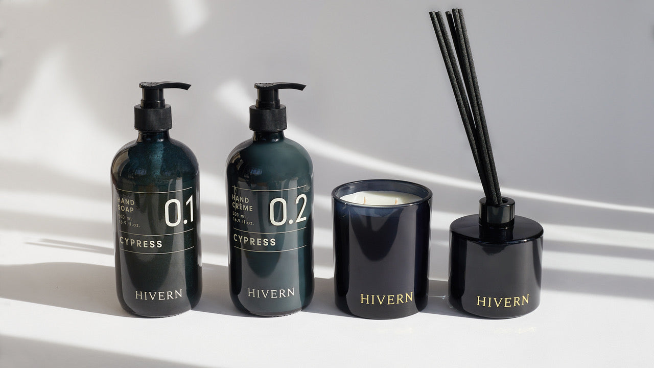 Hivern products lined up order of: Hivern Cypress Hand Soap, Hivern Cypress Hand Crème, Hivern double wicked soya wax candle, Hivern fragrance diffuser. The products sit in front a white background, casting long natural shadows from the light of a nearby window