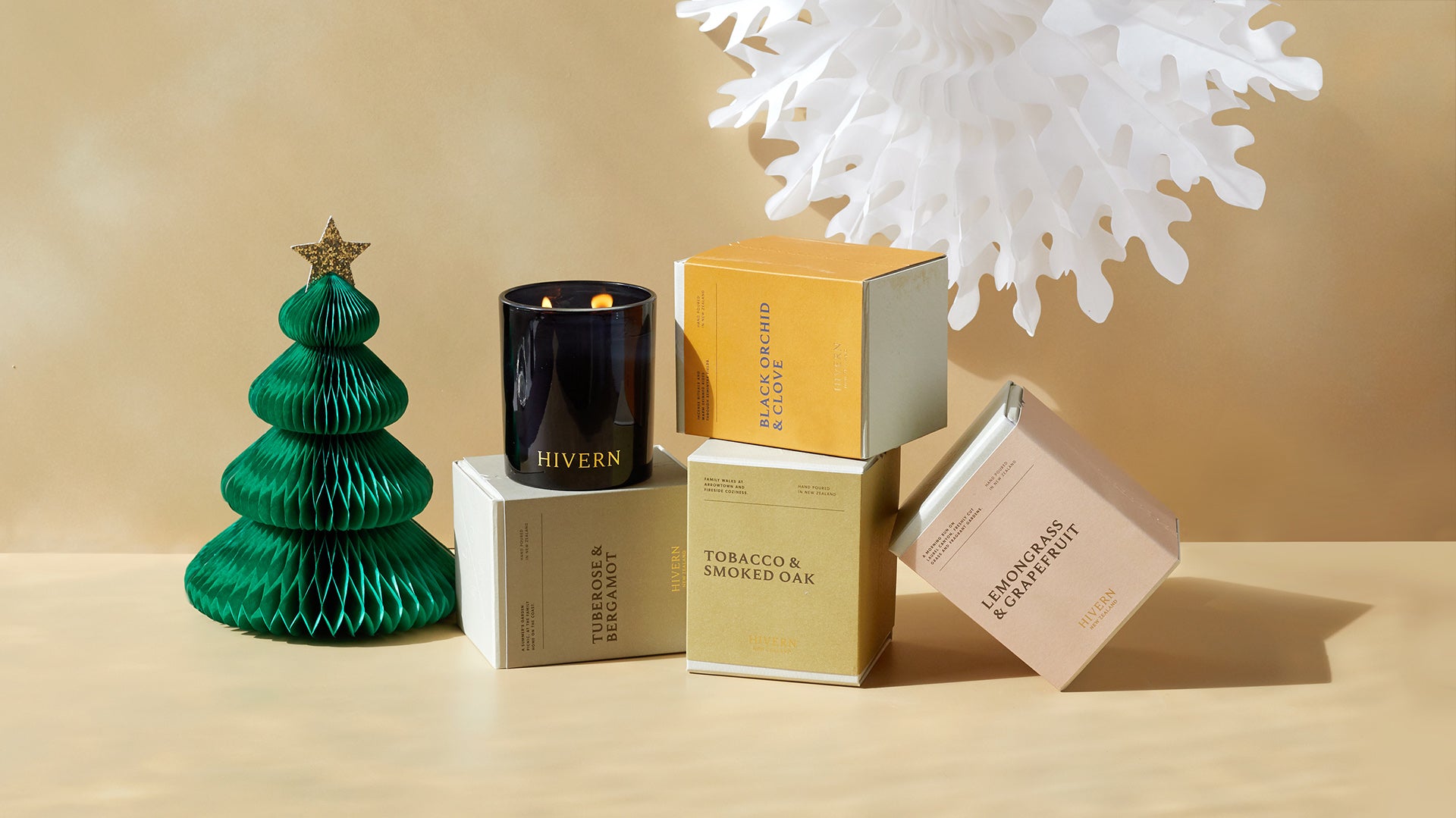 A lit double wicked candle sits amongst colourful packaging and christmas themed decorations against a beige background.