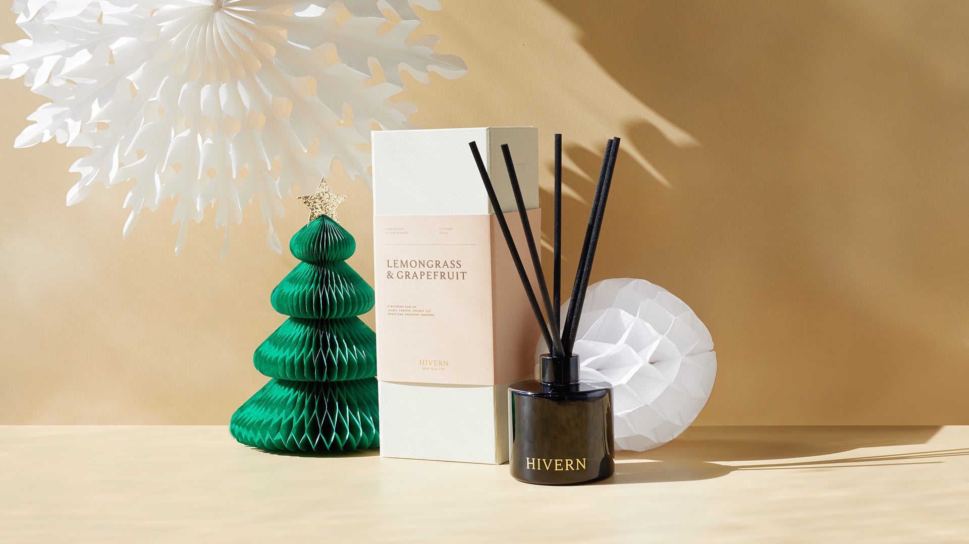 A dark glass fragrance Hivern diffuser with black silk reeds sits next to it's packaging surrounded by Christmas decorations against a beige background.