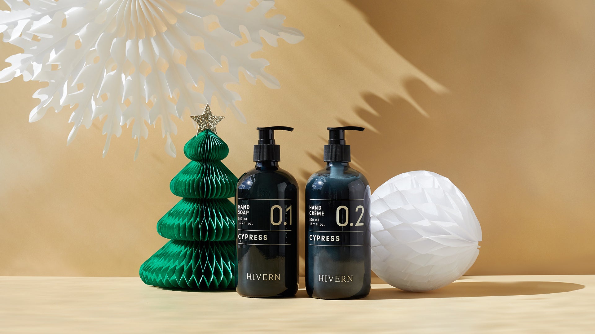 A dark glass pump bottle of Hivern hand soap sits next to a dark glass pump bottle of Hivern hand creme. The bottles are flanked by a green paper Christmas tree decoration and a white paper bauble decoration. The background is beige with a large hanging white paper snowflake deocration.