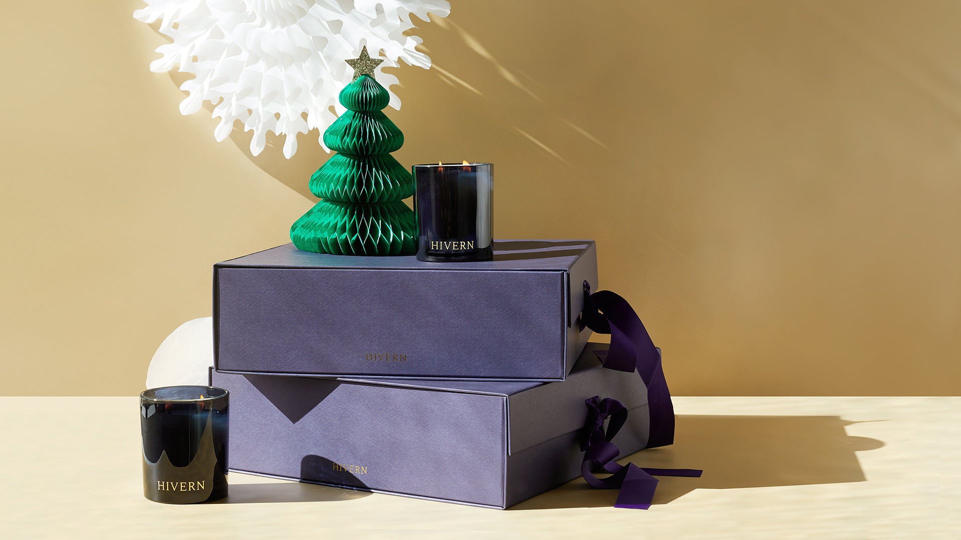 Two lit double wicked candles in dark glass sit next to two stacked purple gift boxes and ribbon with a small paper Christmas tree decoration sitting on top.