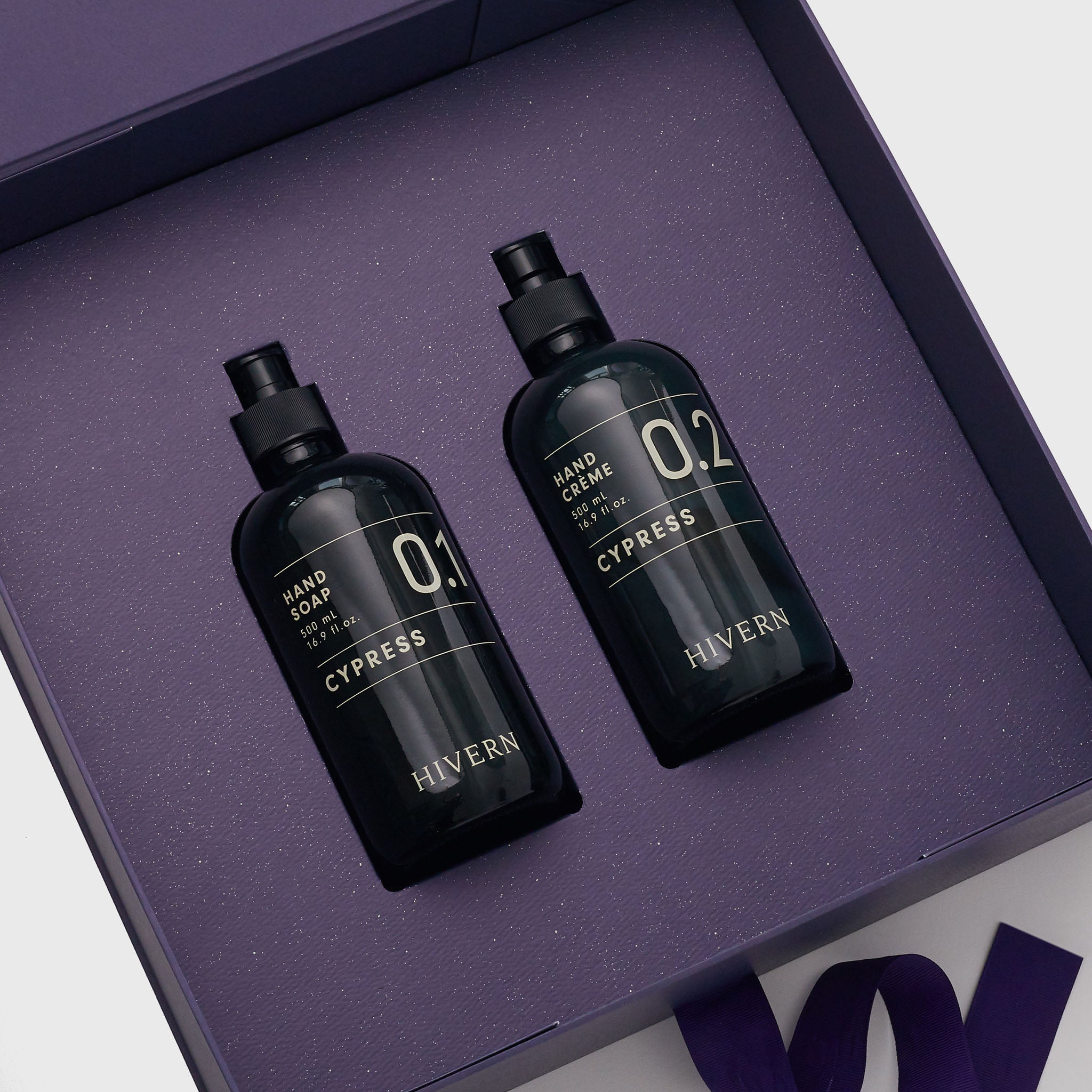 A Hivern luxury giftbox lays open, displaying it's contents. Including a navy blue glass pump bottle of Hivern Cypress Hand Soap and a navy blue glass pump bottle of Hivern Hand Crème.