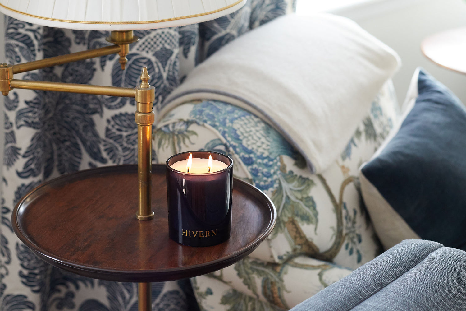 A lit double wicked soya wax candle enclosed in navy blue glass sits on a decorative wooden side table alongside a brass lamp. The table sits next to a sofa chair with detailed nature patterns depicting birds and flora. The sofa chair has a white woolen blanket draped over the top. In the background is a blue and white floral filigree patterned curtain