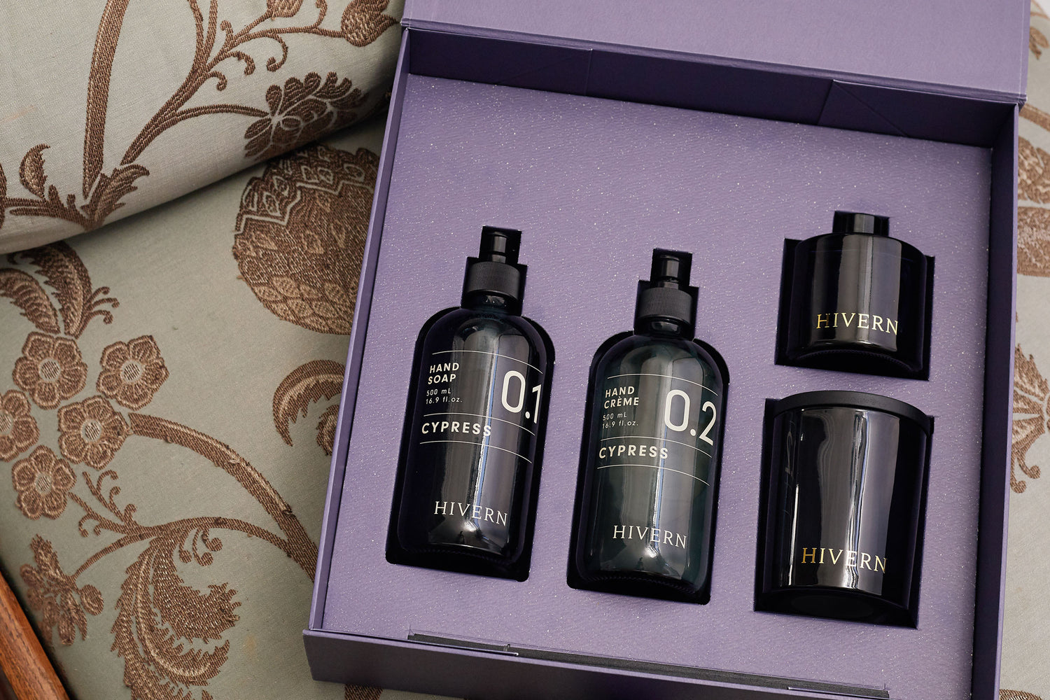 Hivern Giftbox laying open on a luxurious patterned sofa. The giftbox is open, displaying the contents of the gift set including: A glass pump bottle of Hivern Cypress Hand Soap, a glass pump bottle of Hivern Cypress Hand Crème, a navy blue glass Hivern fragrance diffuser and a navy blue glass enclosed Hivern double wick soya wax candle.